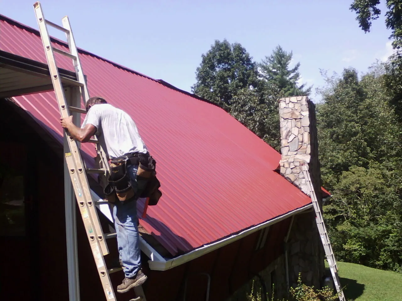 Roofer on a ladder beside a red metal roof and stone chimney — Soffit & Fascia Repair by Roofing Brothers in Allenton